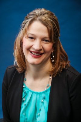 Photo of Jessica Harrell in front of a blue background wearing a black blazer and teal shirt