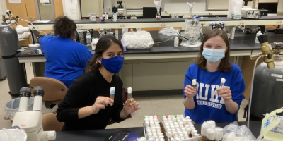 two students working in a socially-distanced lab setting during summer 2021. They are smiling behind their masks, holding test tubes. 